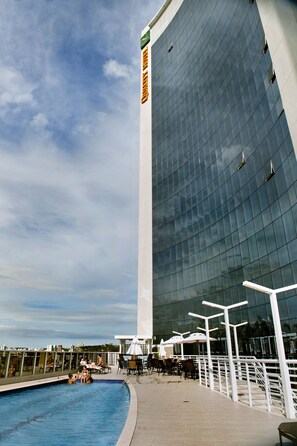A modern building with a reflective glass facade, a pool area with lounge chairs and umbrellas, and a white railing along the pool's edge. - Quality Hotel Vitoria (Vitoria)