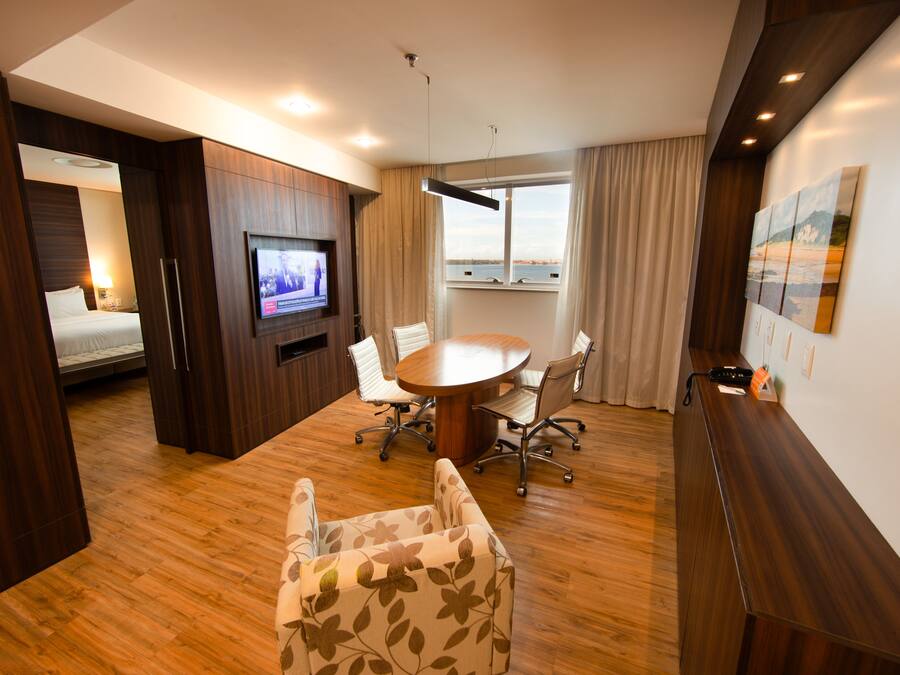 A modern hotel room with a wooden floor, a round dining table, a TV, a bed, and a large window with a view.