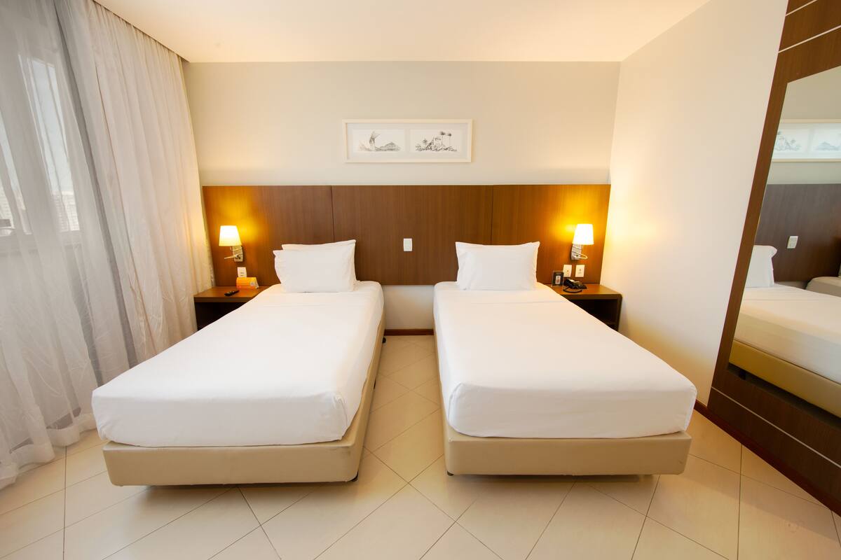 A hotel room with two single beds, a wooden headboard, bedside lamps, a framed artwork on the wall, and a large mirror.