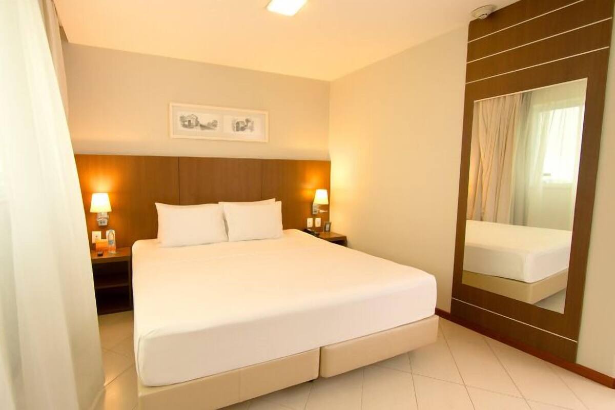 A hotel room with a large bed, two bedside tables with lamps, a headboard, a mirror, and a framed picture on the wall.
