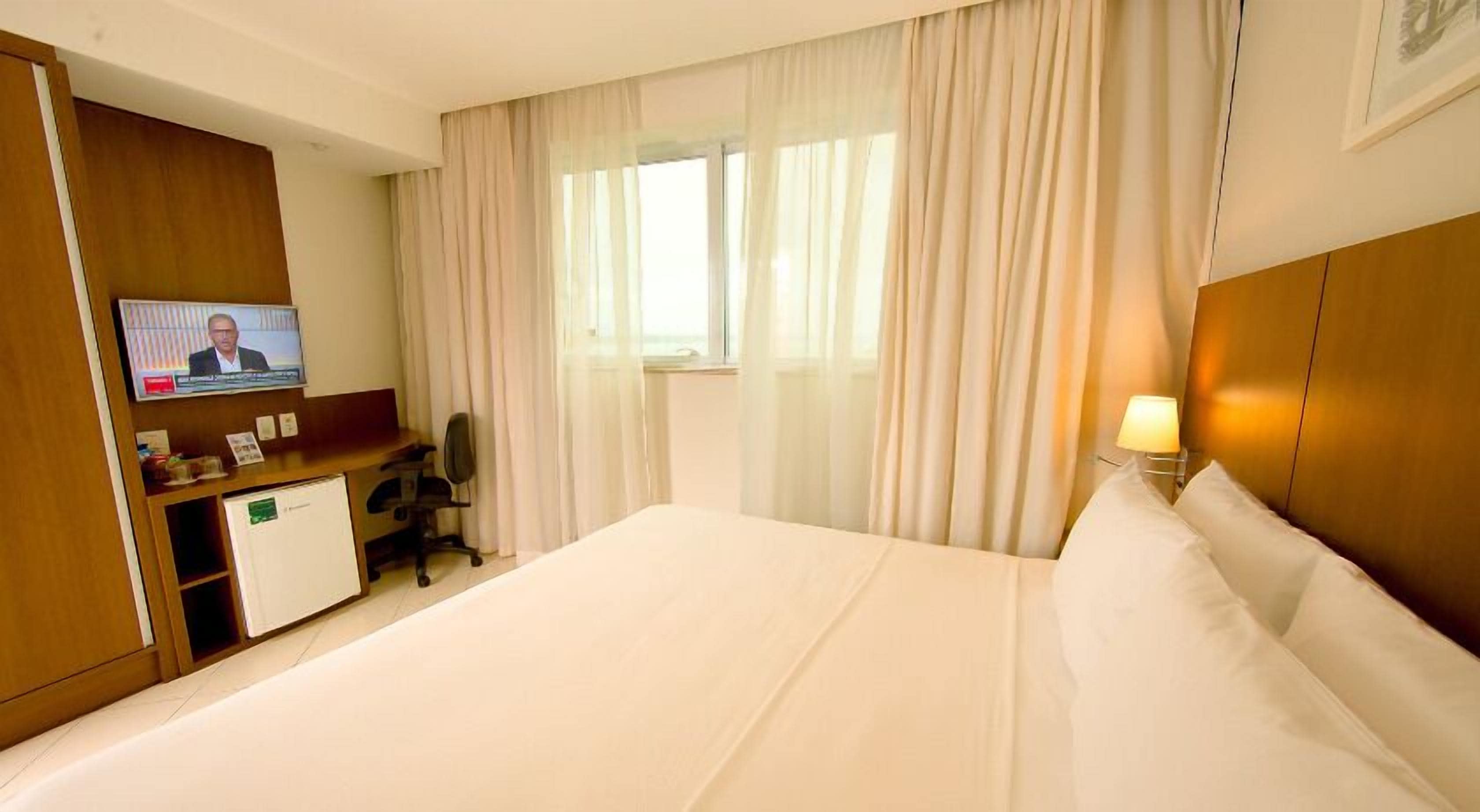 Quarto de hotel com uma cama grande, uma escrivaninha com computador, televisão e janela com cortinas.