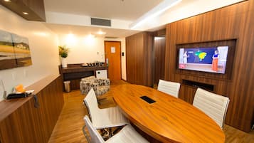 Uma sala de conferências moderna com uma mesa de madeira, cadeiras brancas, uma tela de TV e um balcão de recepção.