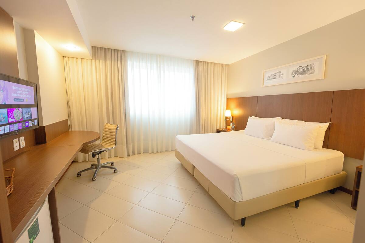 A hotel room with a large bed, a desk with a computer, a chair, and a window with curtains.