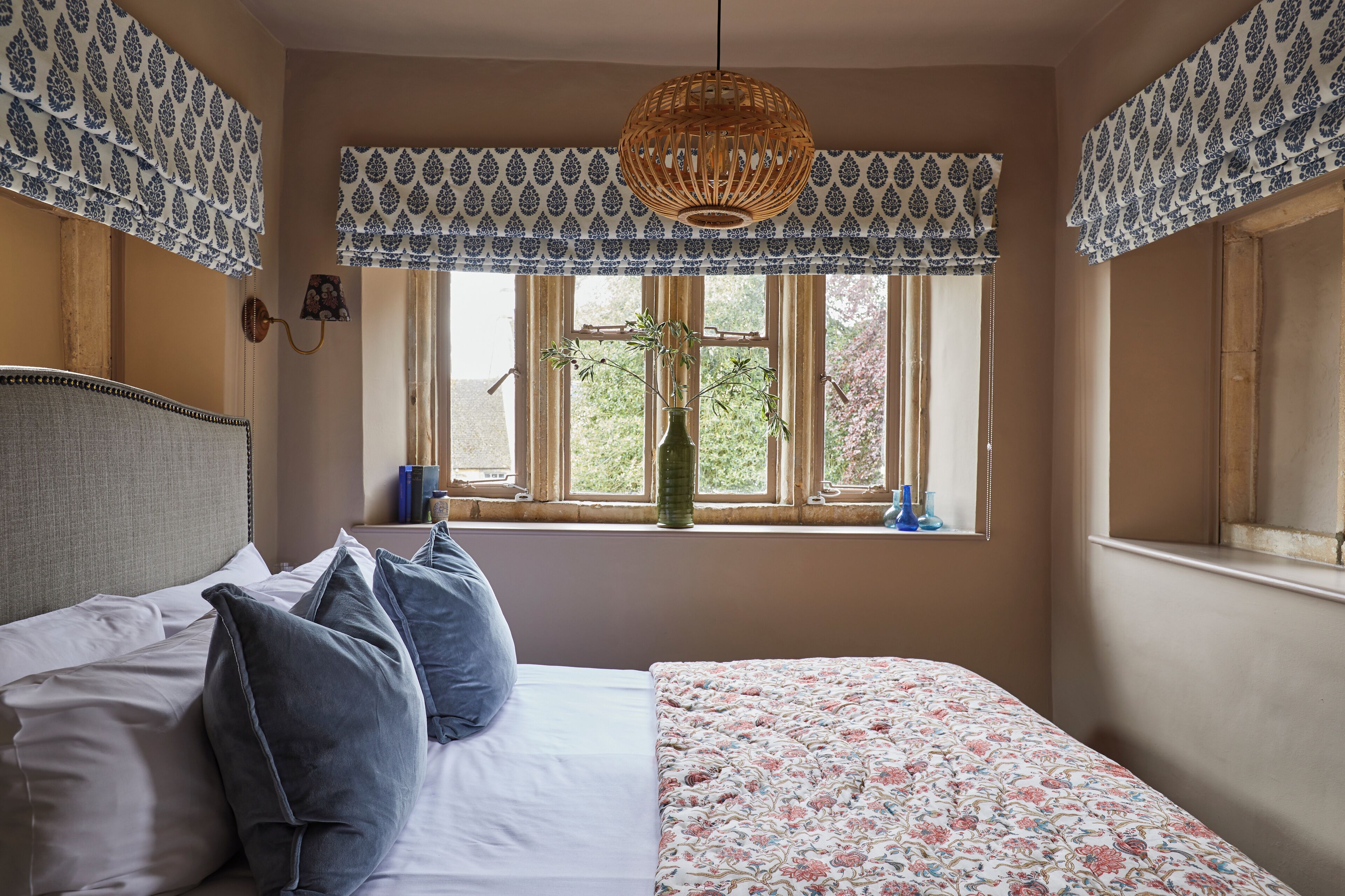 Un dormitorio con cama, almohadas, una ventana con cortinas, una lámpara colgante y un florero con plantas.