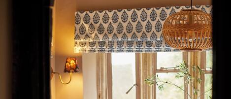 Ein Schlafzimmer mit Bett, Kissen, einem Fenster mit Vorhängen, einer Pendelleuchte und einer Pflanze.