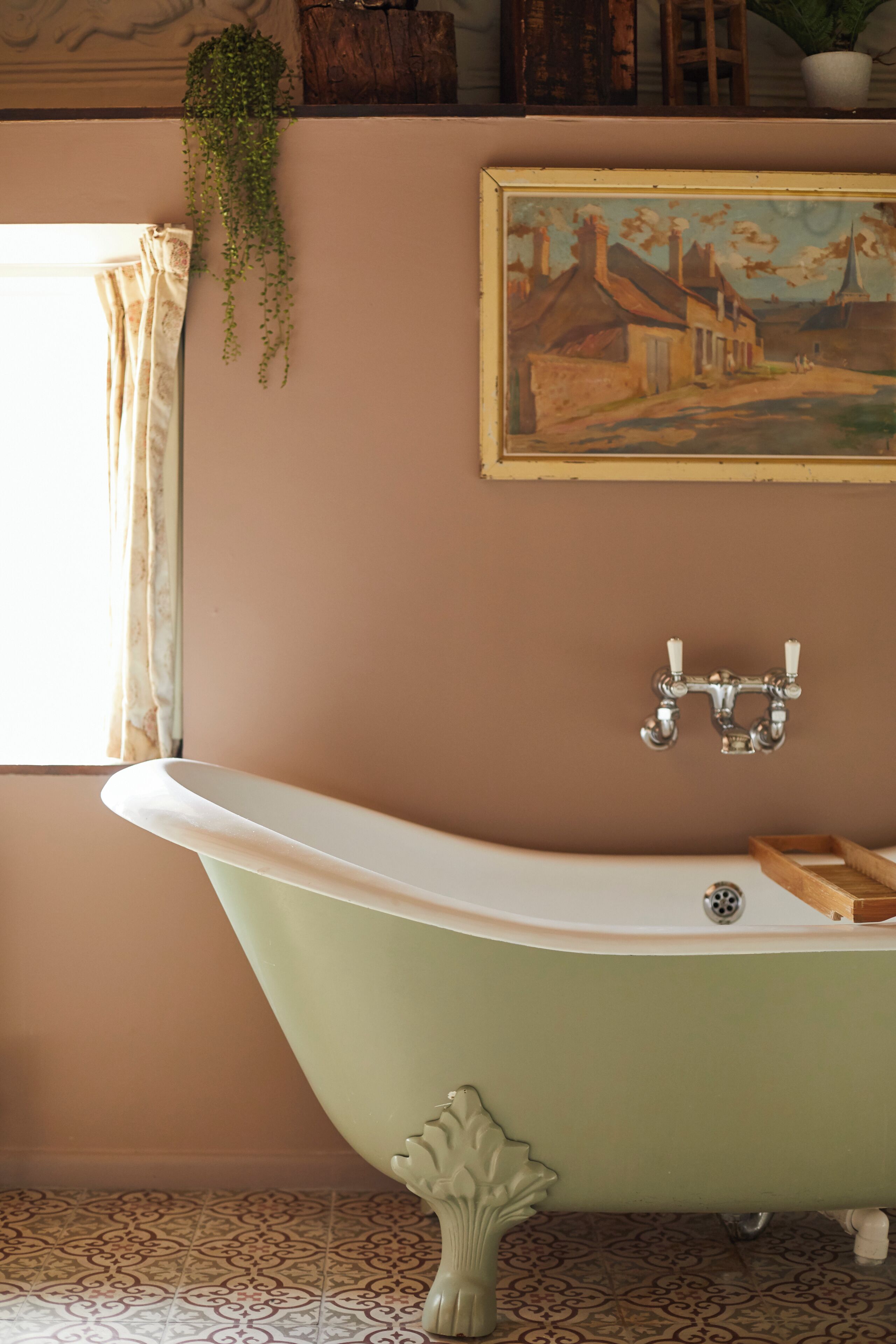 A bathroom with a freestanding bathtub, a window with curtains, a potted plant, and a painting on the wall.
