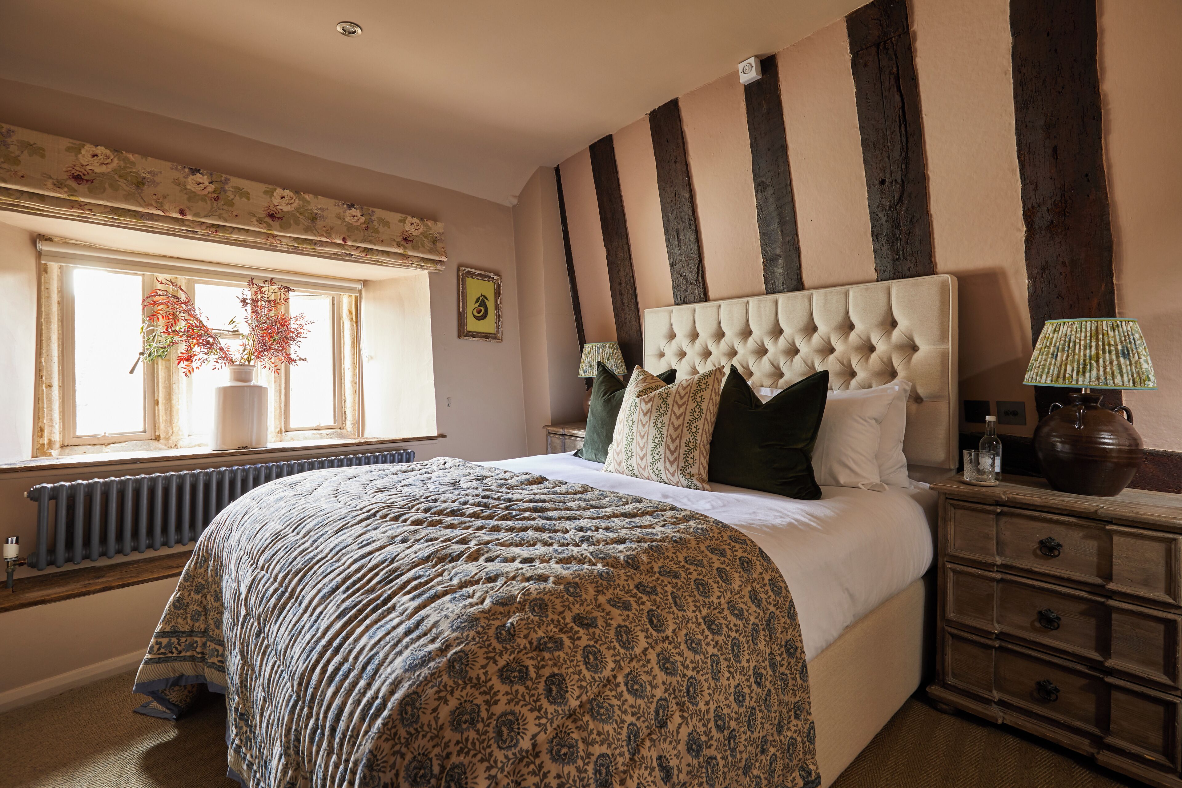 A bedroom with a large bed, a window with curtains, a radiator, a wooden dresser, and a lamp.