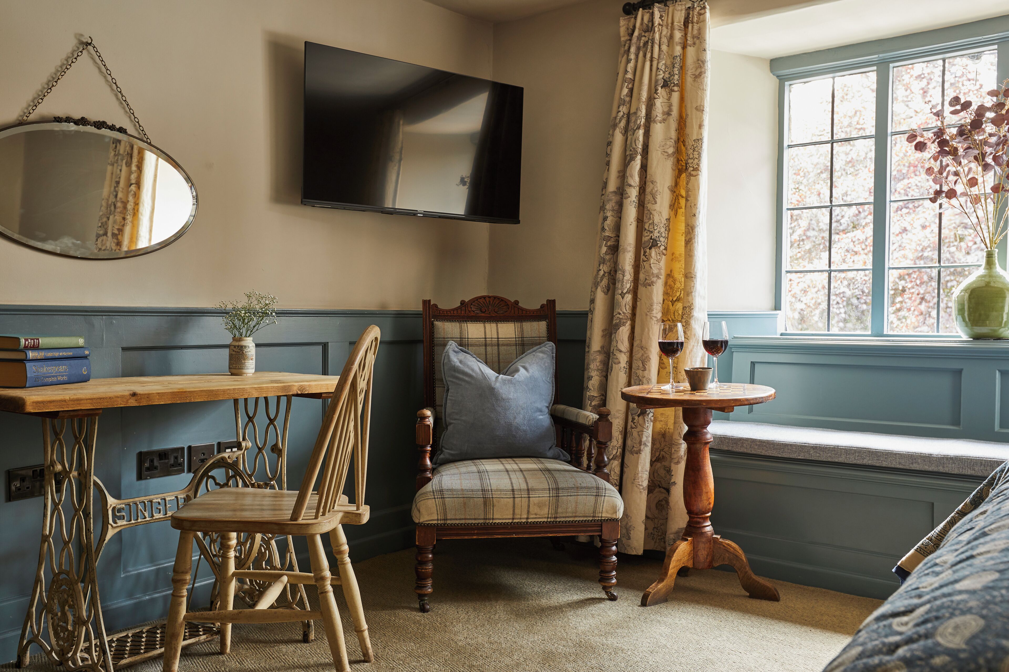 A cozy room with a wooden chair, a small table with a vase and wine glasses, a television mounted on the wall, and a window with a view of trees.