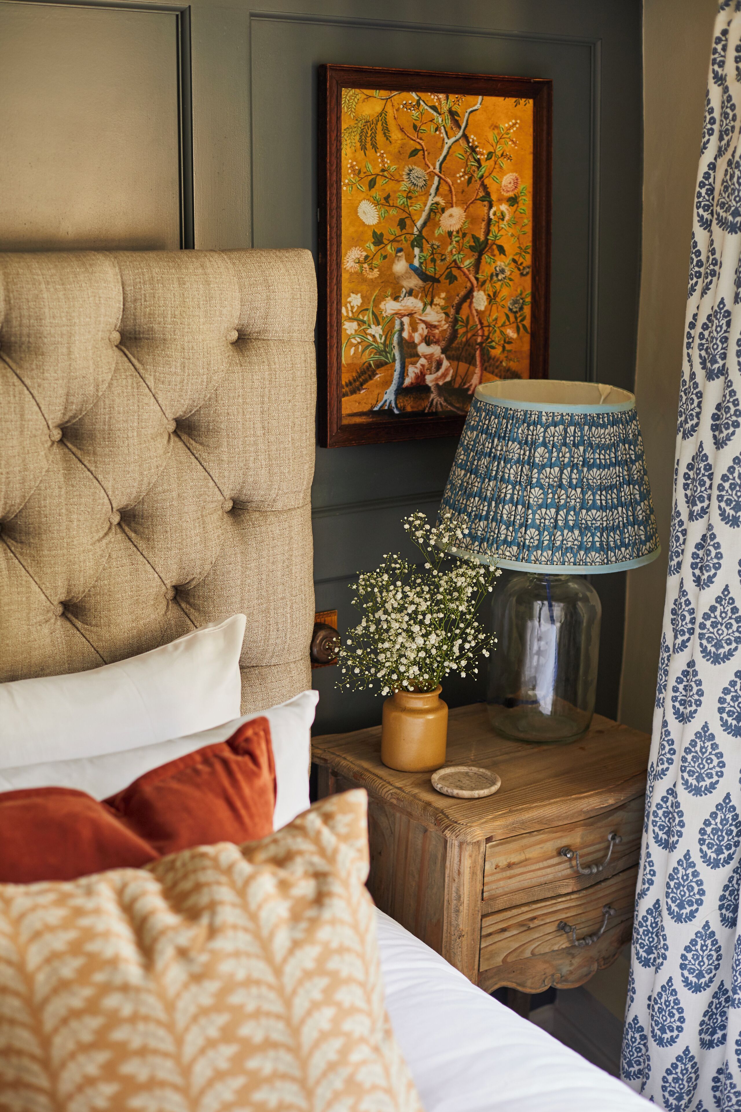 Une chambre à coucher avec une tête de lit capitonnée, un tableau, une lampe de chevet, un vase de fleurs et un rideau à motifs.