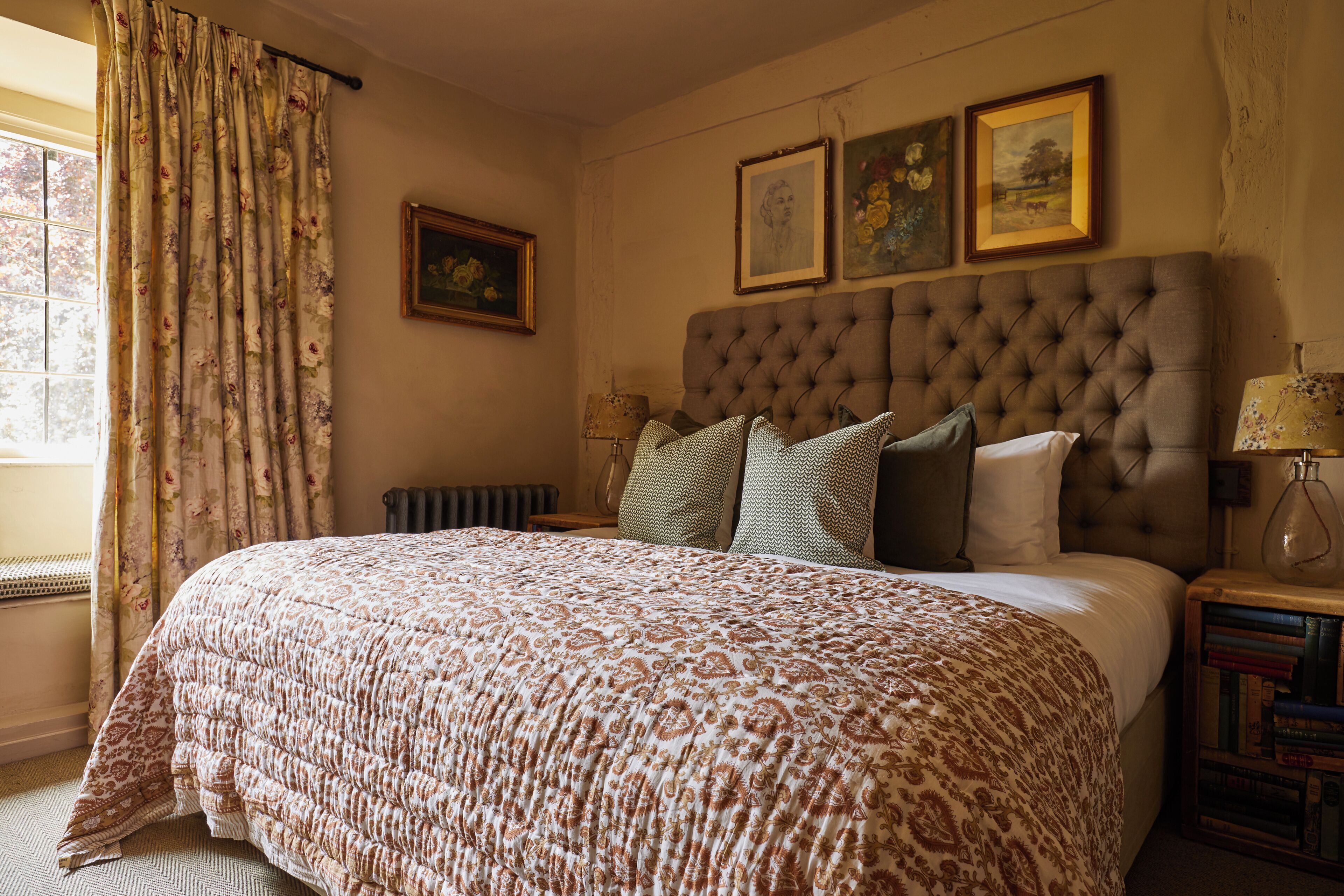 A bedroom with a large bed, a radiator, a window with curtains, and a bedside table with books.