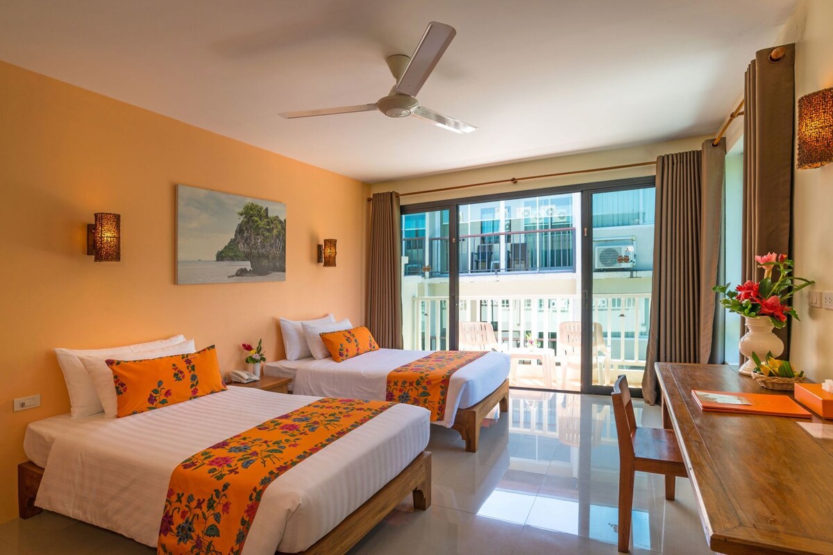 A hotel room with two beds, a balcony, a dining table, and a ceiling fan.