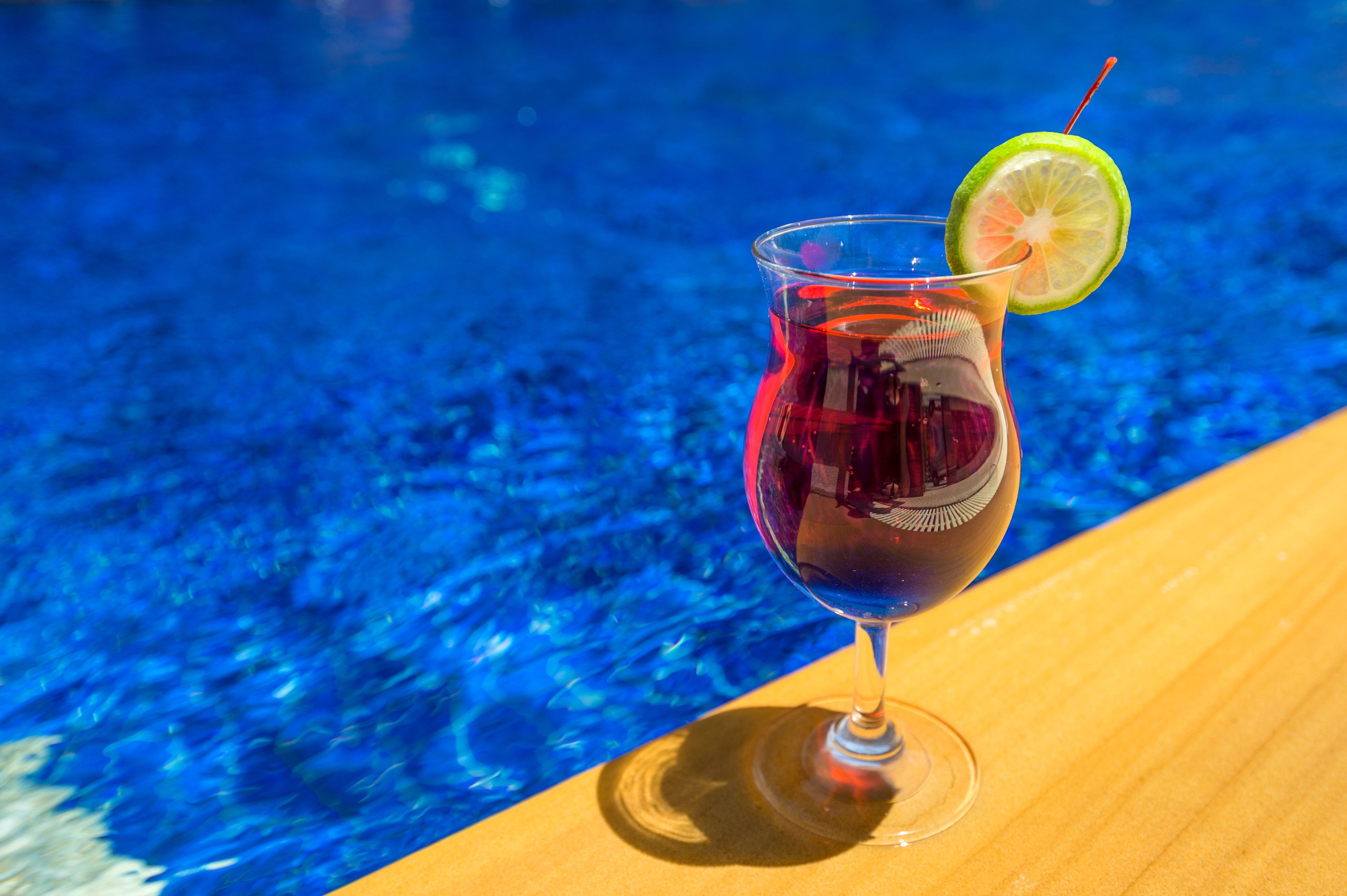 A glass of red drink with a lime garnish on a wooden surface by a pool.