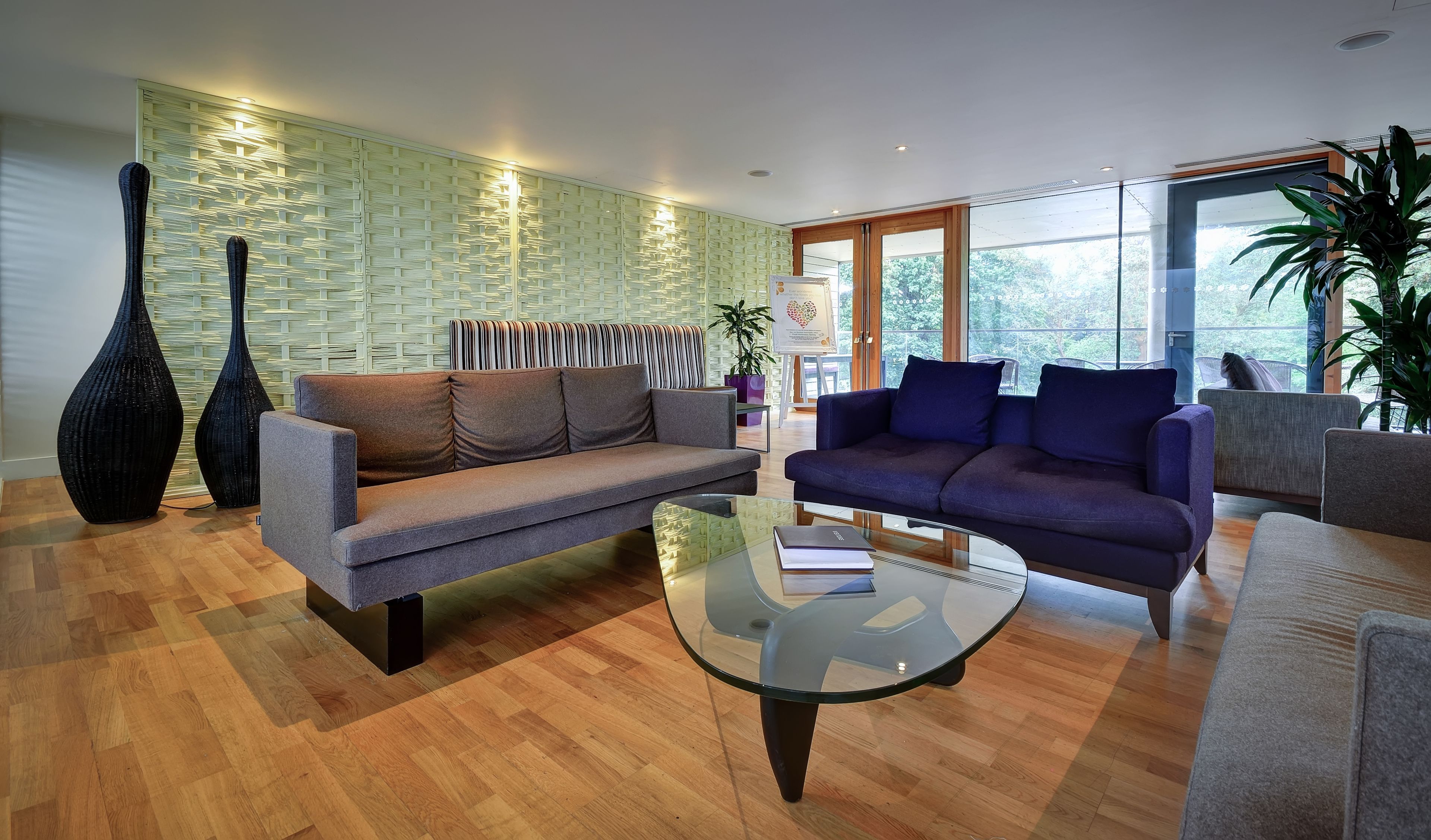 A modern living room with a grey sofa, a blue sofa, and a glass coffee table. There are two large vases and a potted plant. The room has wooden flooring and a large window with a view of trees.