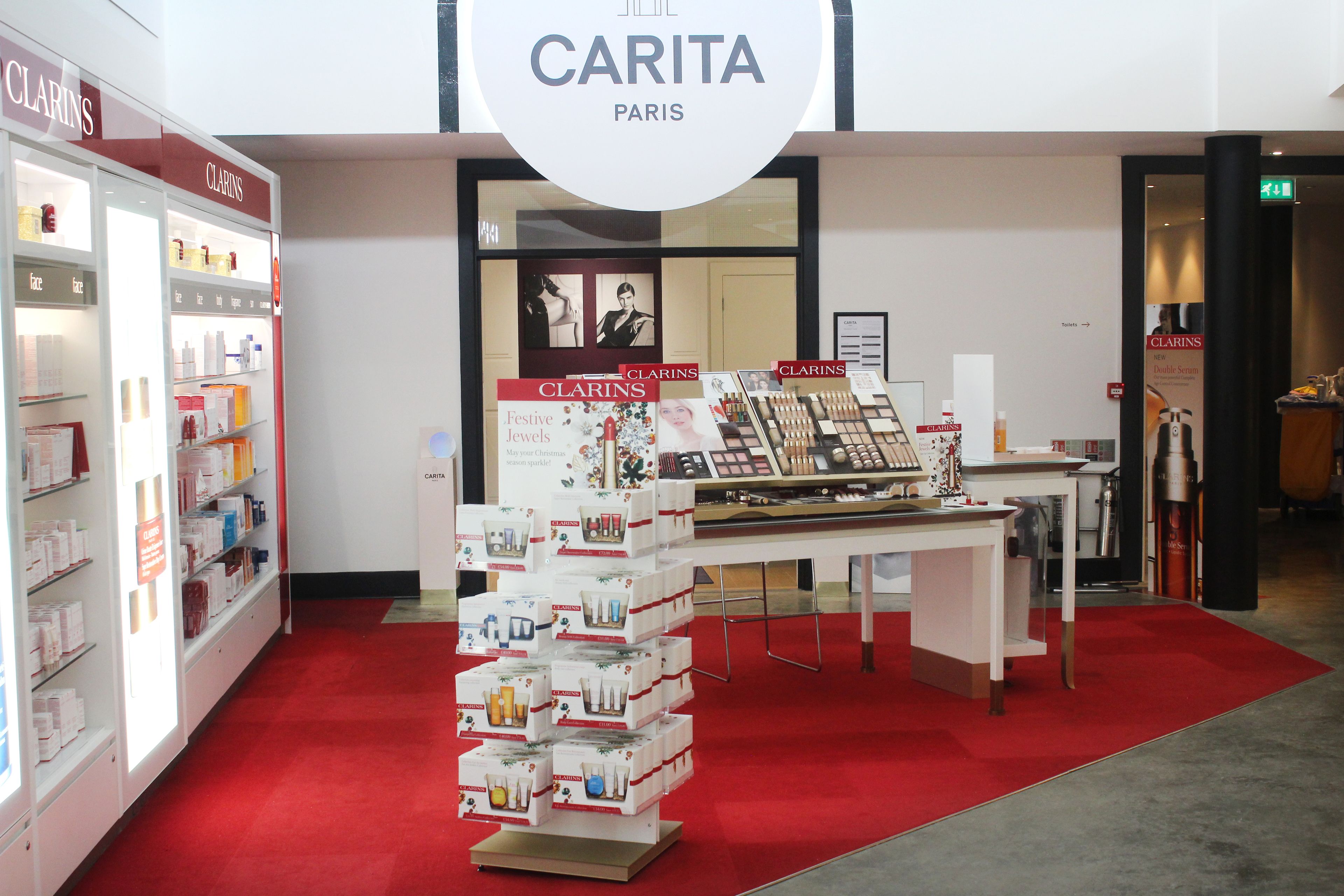Een winkelinterieur met een rode loper, een displaytafel met diverse producten, en een bordje dat 'CARITA PARIS' vermeldt.