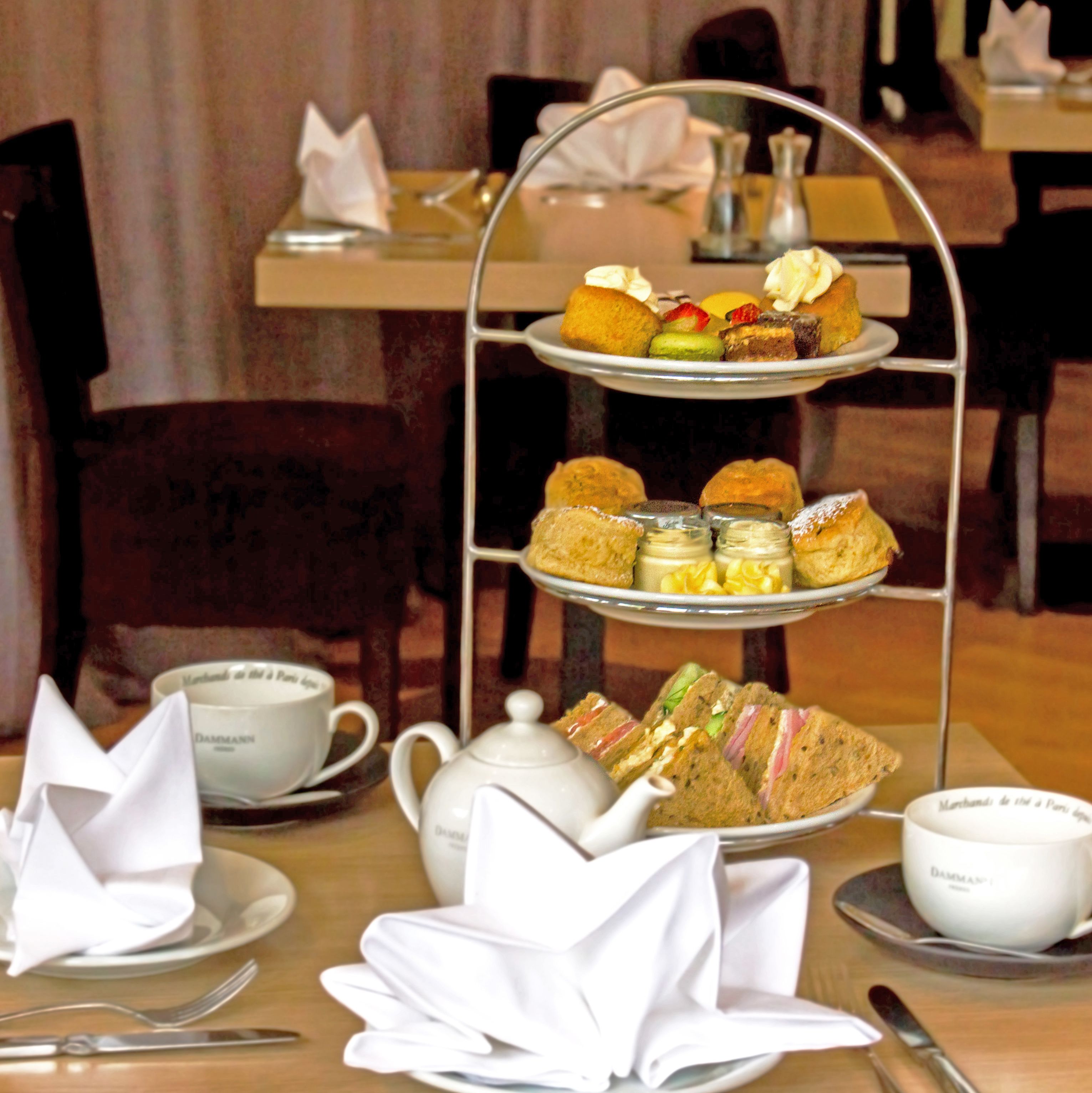 A three-tiered dessert stand with various pastries and sandwiches, a teapot, cups, and cutlery on a table.