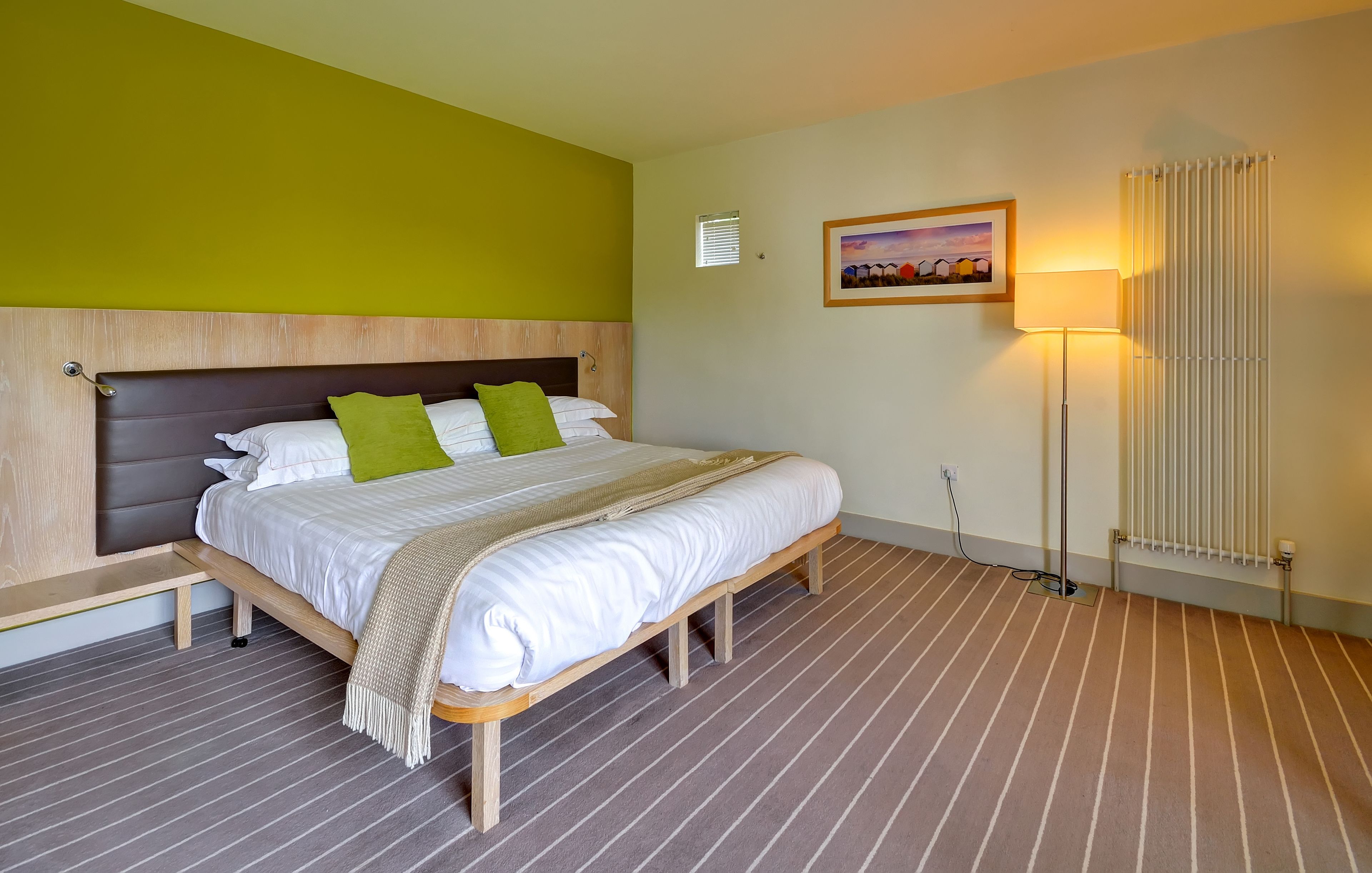 Una cama bien hecha con cabecera de madera, una mesita de noche, una lámpara de pie y un radiador.