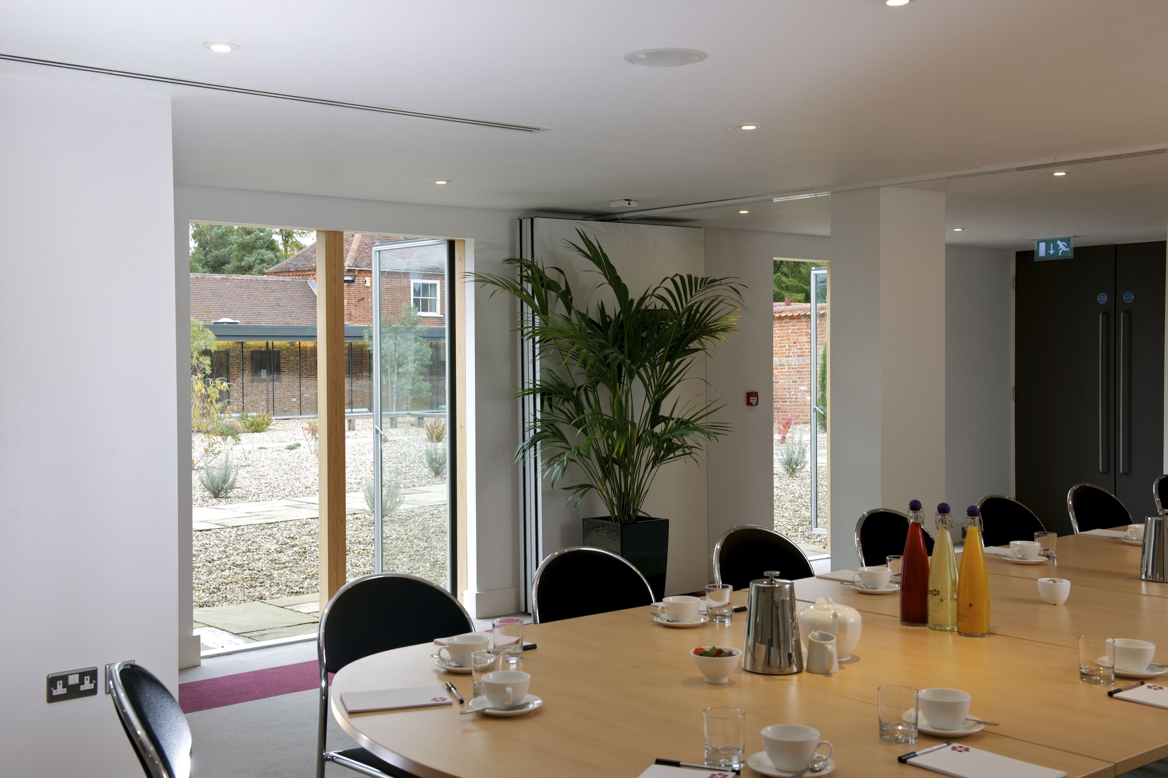 A modern conference room with a large table set for a meeting, surrounded by chairs, and a large window offering a view of the outdoors.