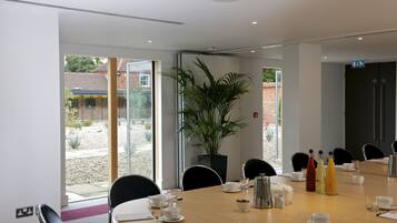 A modern conference room with a large table set for a meeting, surrounded by chairs, and a large window offering a view of the outdoors.