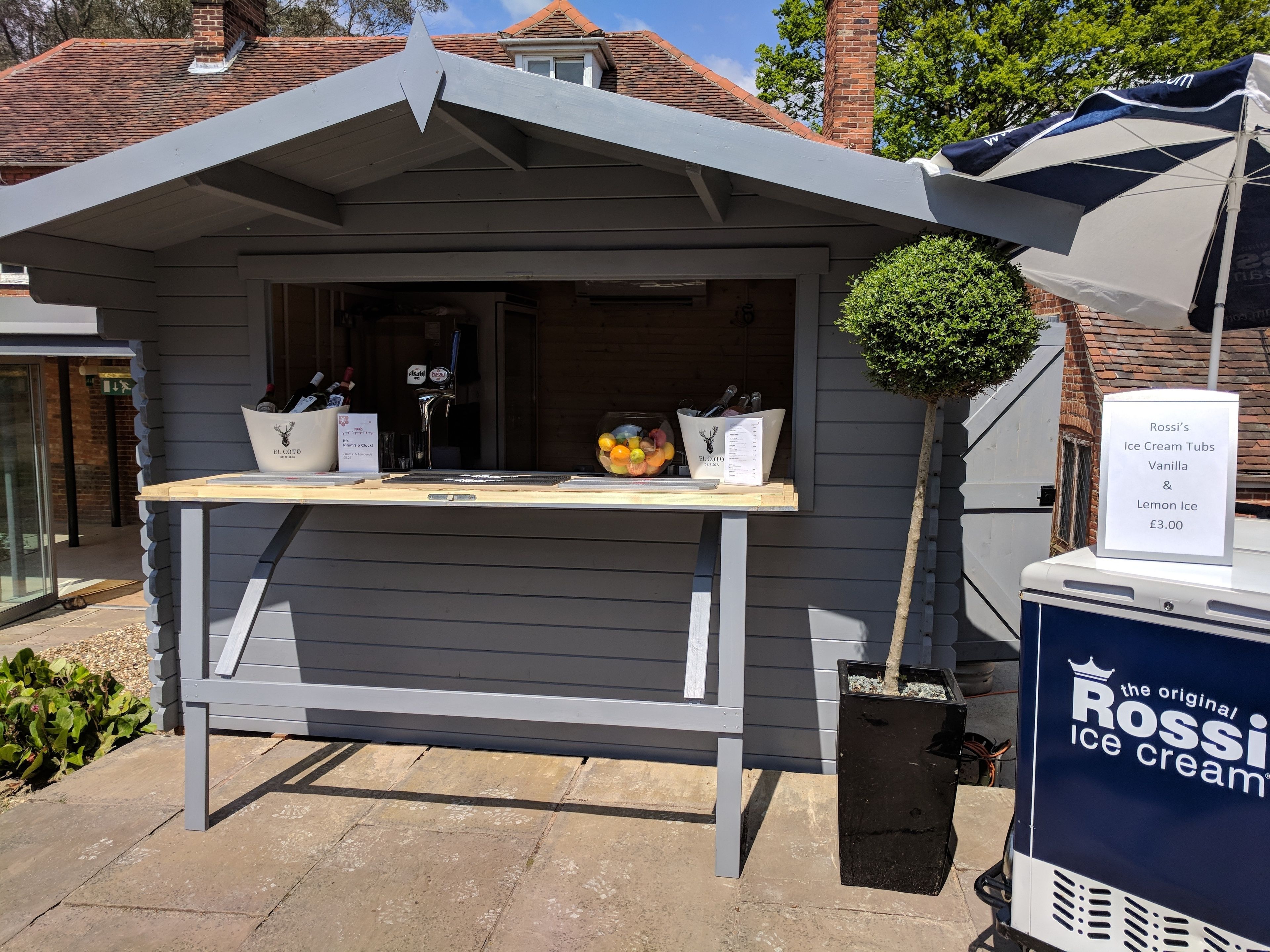 A small outdoor ice cream stand with a sign advertising Rossi's Ice Cream Tubs Vanilla & Lemon Ice for £3.00.