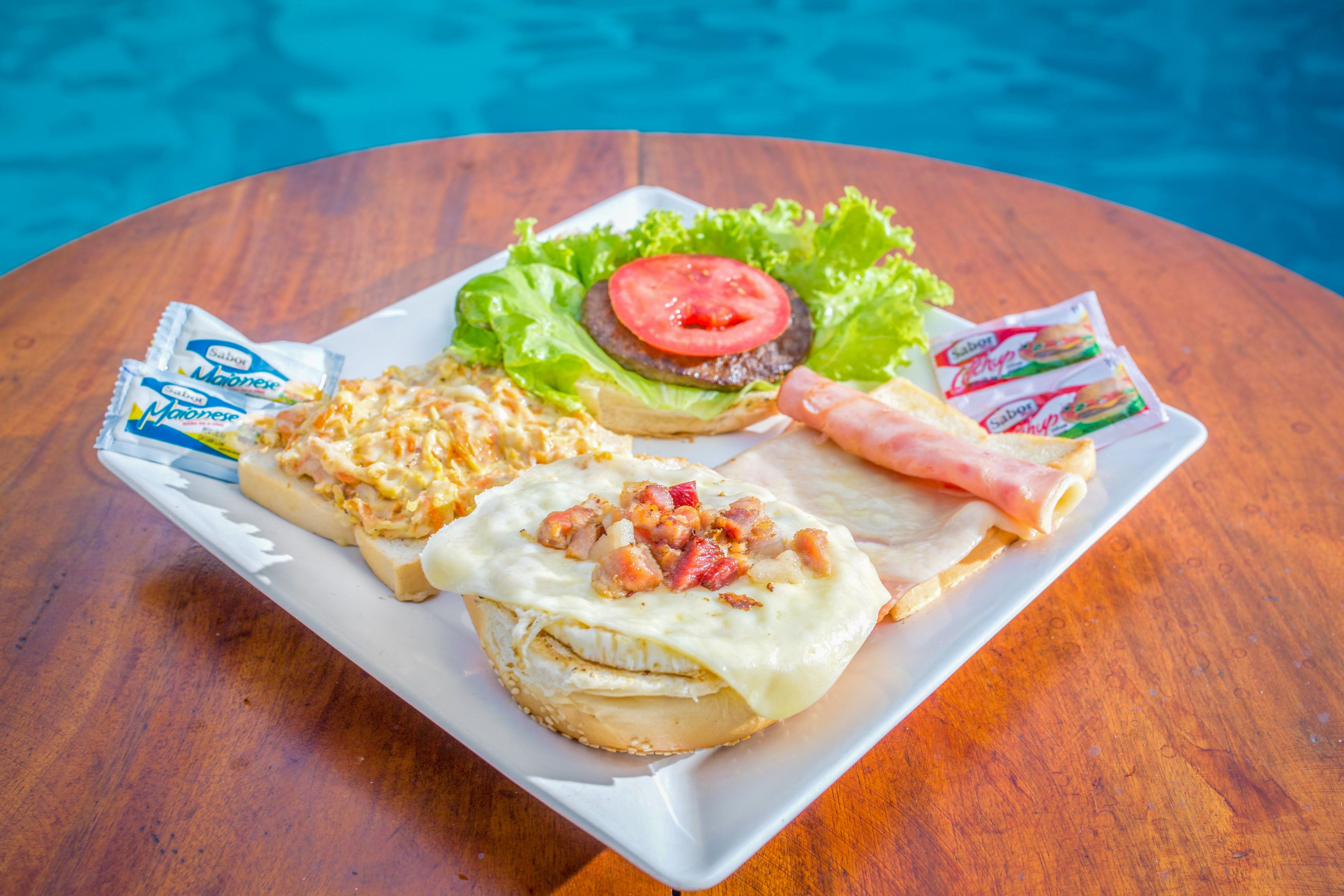 A plate with a sandwich, salad, and condiments on a wooden table by a pool.