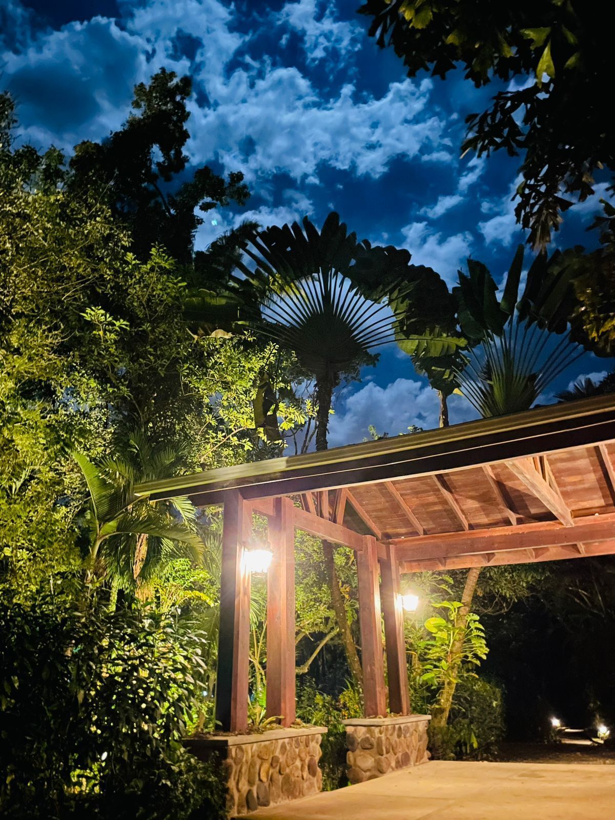 A wooden gazebo with stone pillars, surrounded by lush tropical vegetation and palm trees.