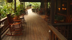 A wooden deck with rocking chairs and tables, surrounded by lush greenery.