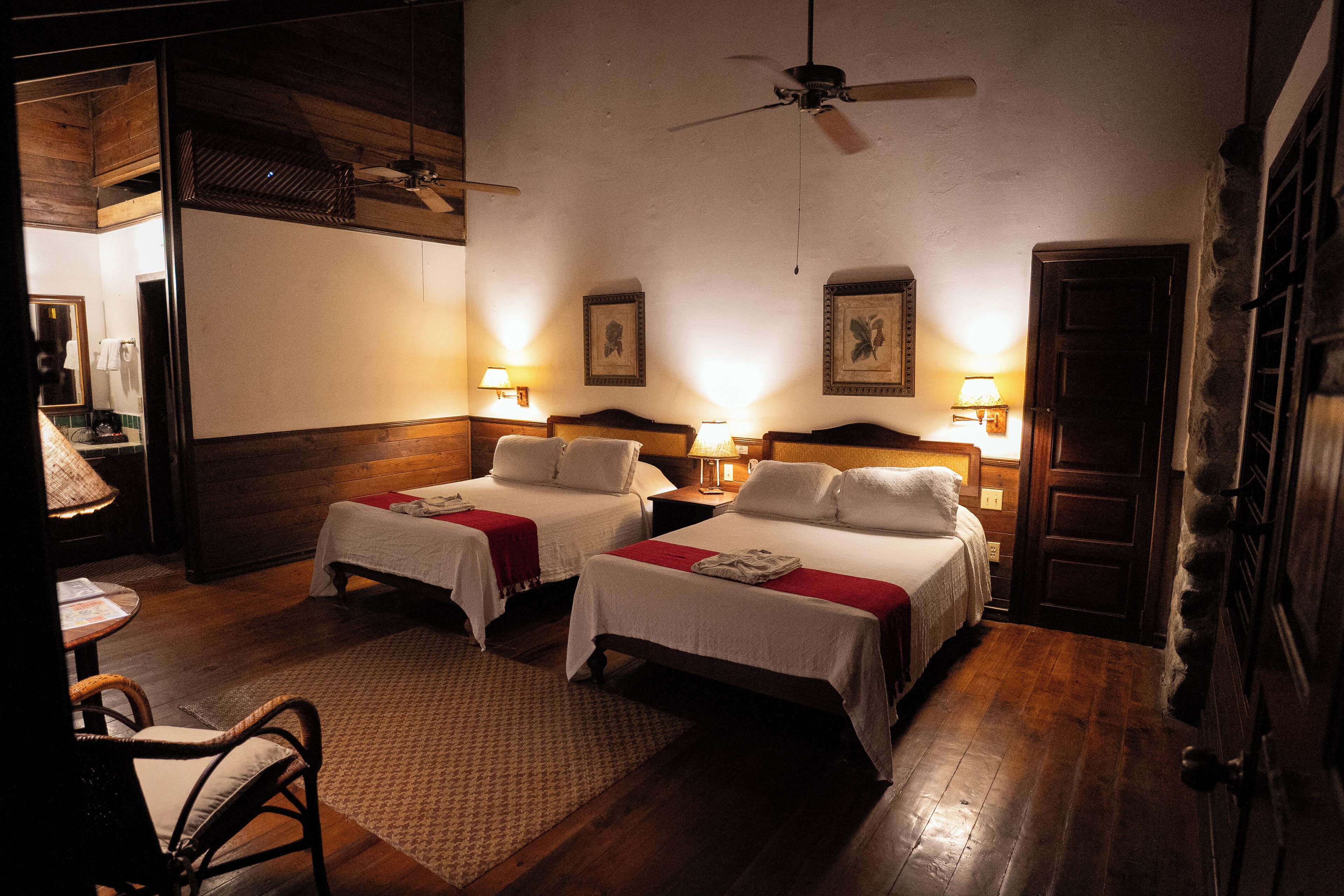 Habitación de hotel con dos camas, piso de madera, ventilador de techo y lámparas de pared.