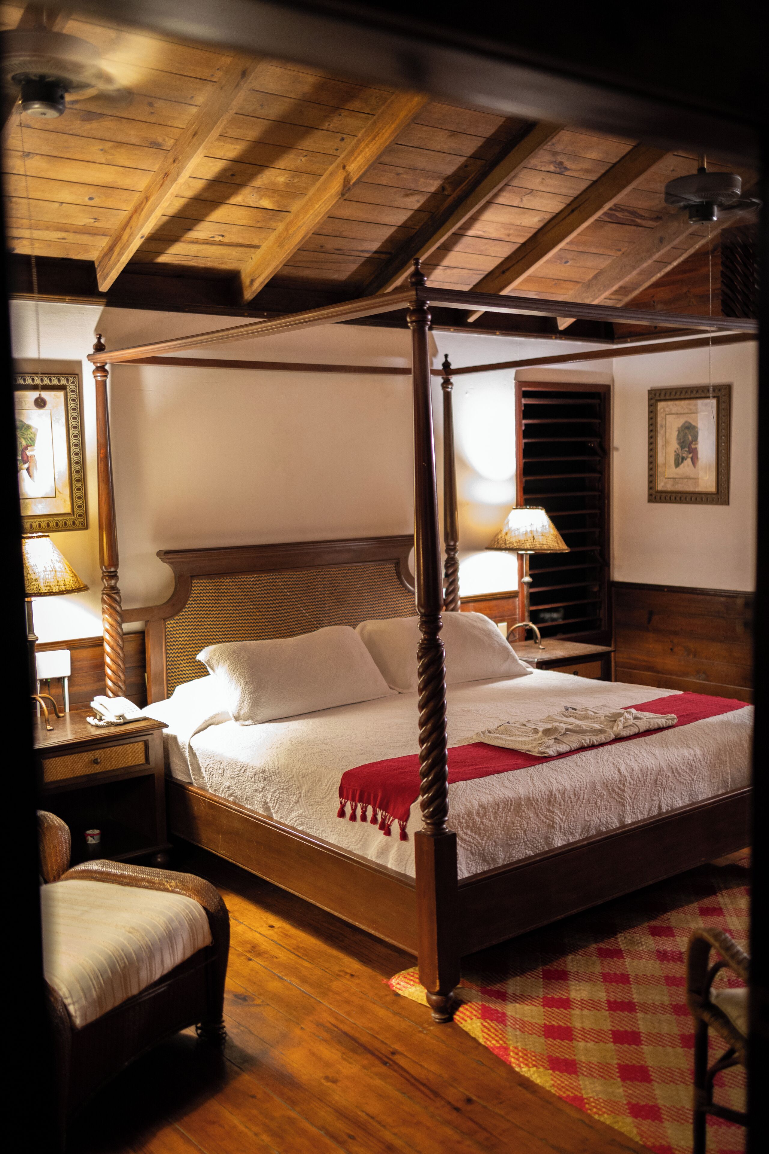 Een hemelbed met een houten hoofdeinde, een rode plaid en een geruit tapijt op de vloer. Er staat een nachtkastje met een lamp en een telefoon erop.
