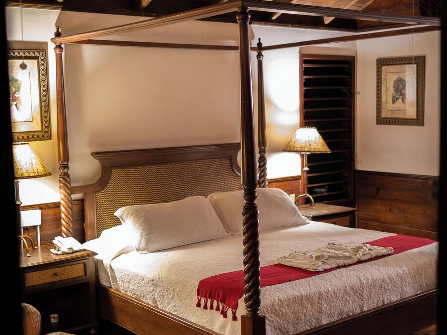Een hemelbed met een houten hoofdeinde, een rode plaid en een geruit tapijt op de vloer. Er staat een nachtkastje met een lamp en een telefoon erop.