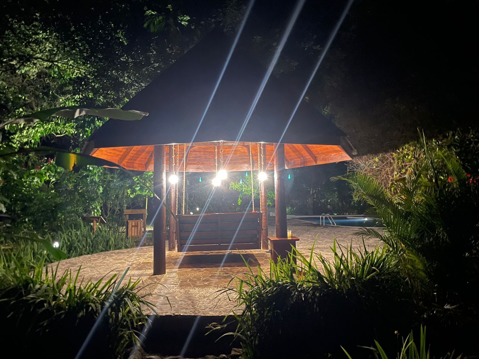 A wooden gazebo with a roof, surrounded by lush greenery and a swimming pool.
