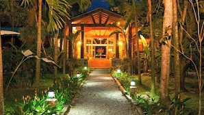 A well-lit pathway leading to a wooden structure with a porch, surrounded by lush greenery and palm trees.