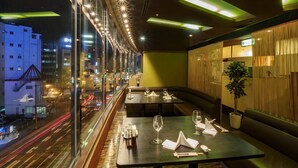 A restaurant with tables and booths, a view of the cityscape, and a green wall with a plant. - Airline Hotel (Miyazaki)