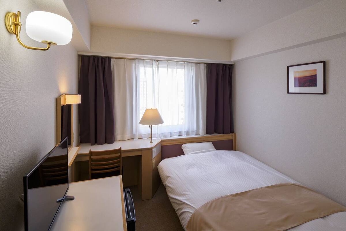 A hotel room with a bed, a desk, a chair, a lamp, a window with curtains, and a picture on the wall.
