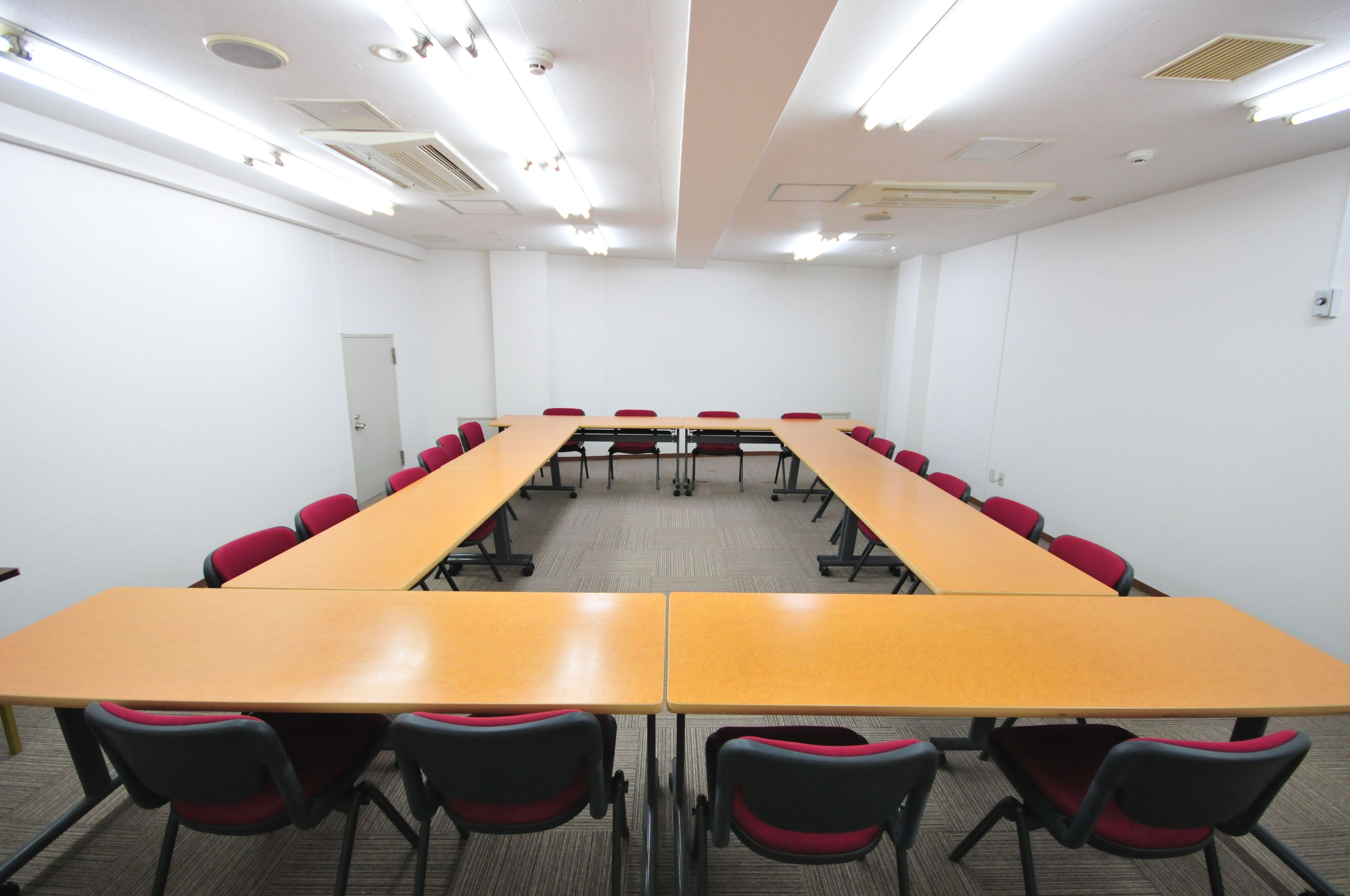 A conference room with a long table and red chairs, white walls, and ceiling lights.