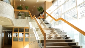 A modern hotel lobby with a marble reception desk, a staircase with patterned steps, and large windows allowing natural light. - Hotel Premium Green Hills (Sendai)