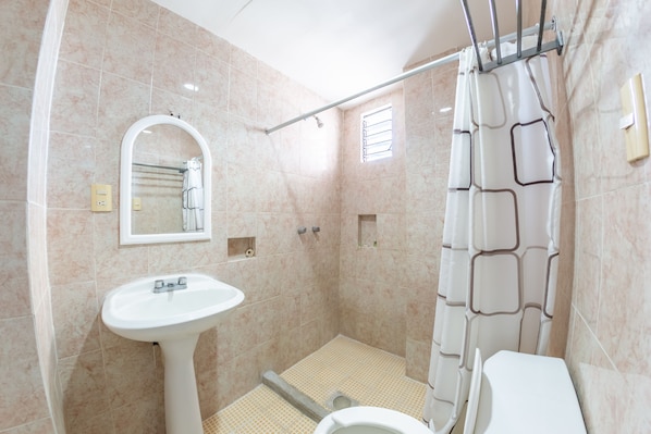 Un baño con lavamanos pedestal, ducha con cortina, ventanal pequeño y espejo.