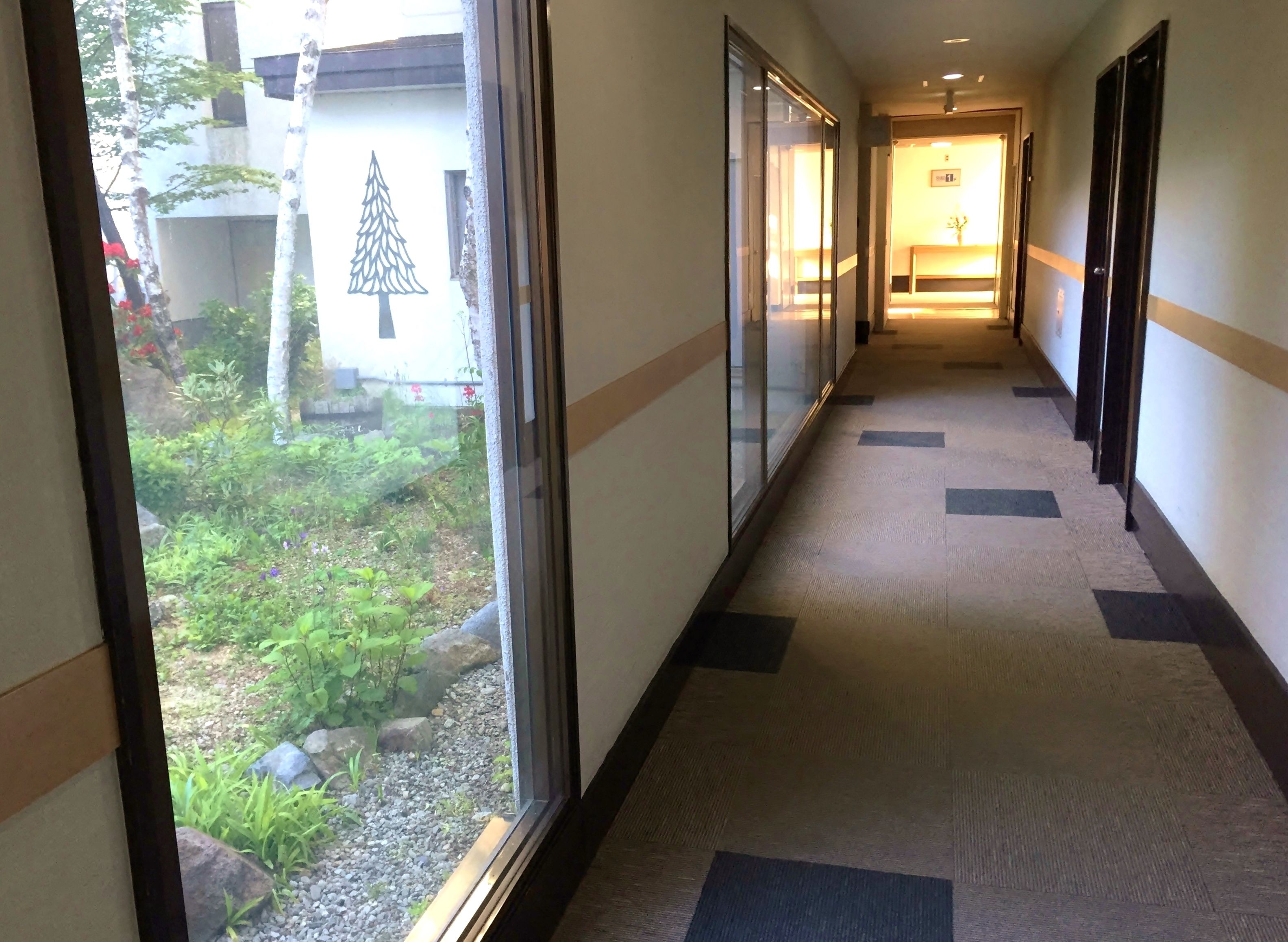 A hotel corridor with a view of a garden outside, a reception area at the end, and a patterned carpet.