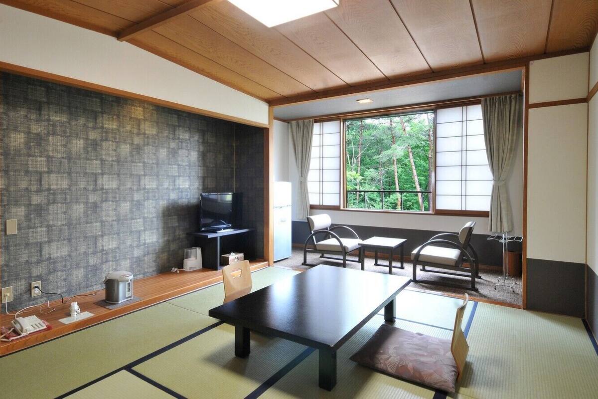 Una habitación japonesa tradicional con suelo de tatami, una mesa baja, sillas, televisión y vista a áreas verdes.