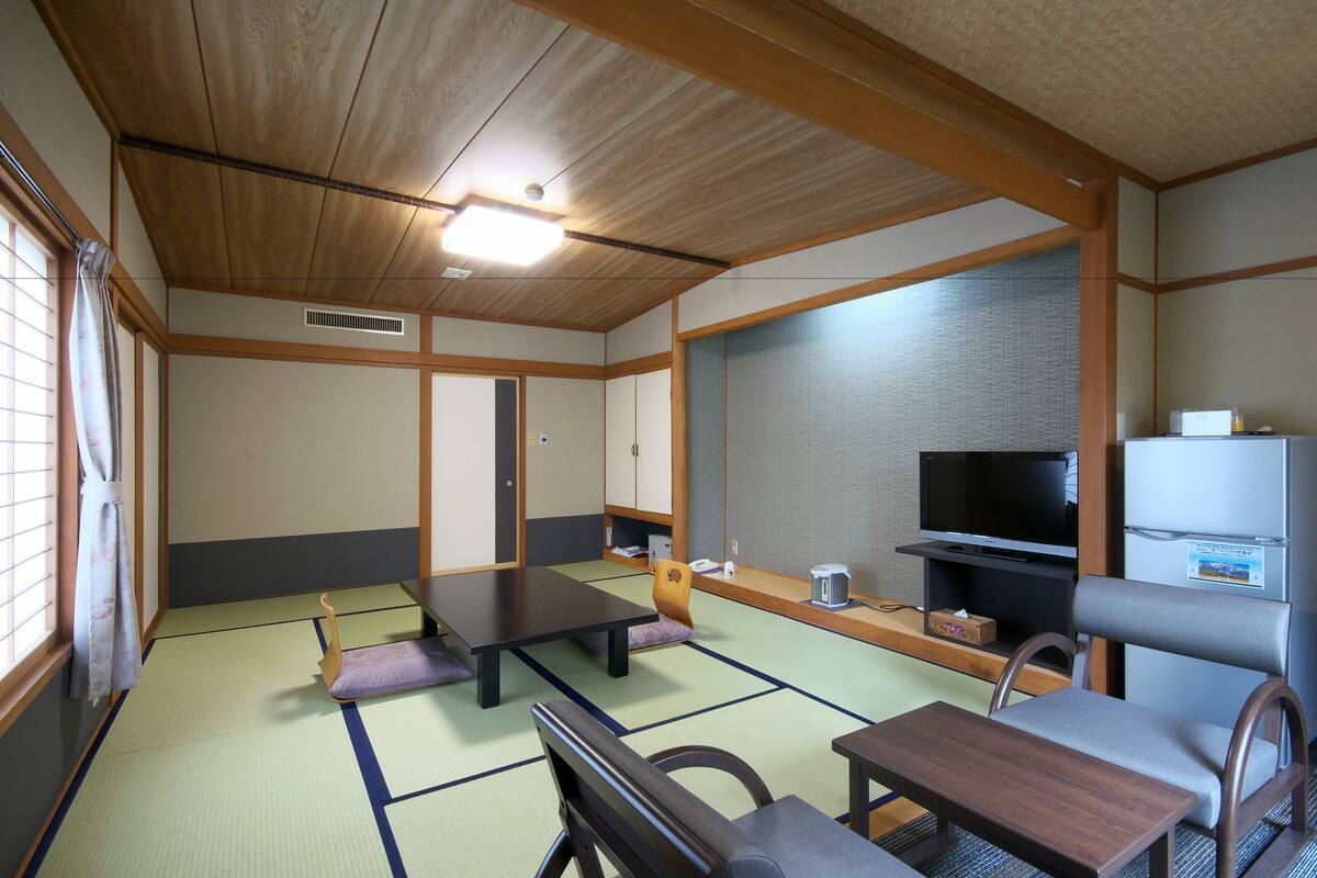 A traditional Japanese-style room with tatami flooring, a low table, a cushioned chair, a television, and a refrigerator.