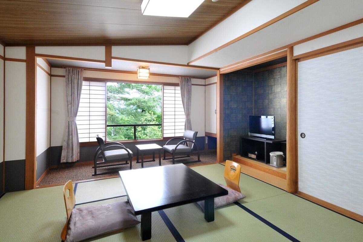 A traditional Japanese room with tatami flooring, a low table, chairs, a television, and a view of greenery outside.