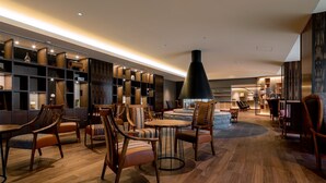 A modern lobby with wooden flooring, a central seating area with round tables and chairs, and a large black pendant light.
