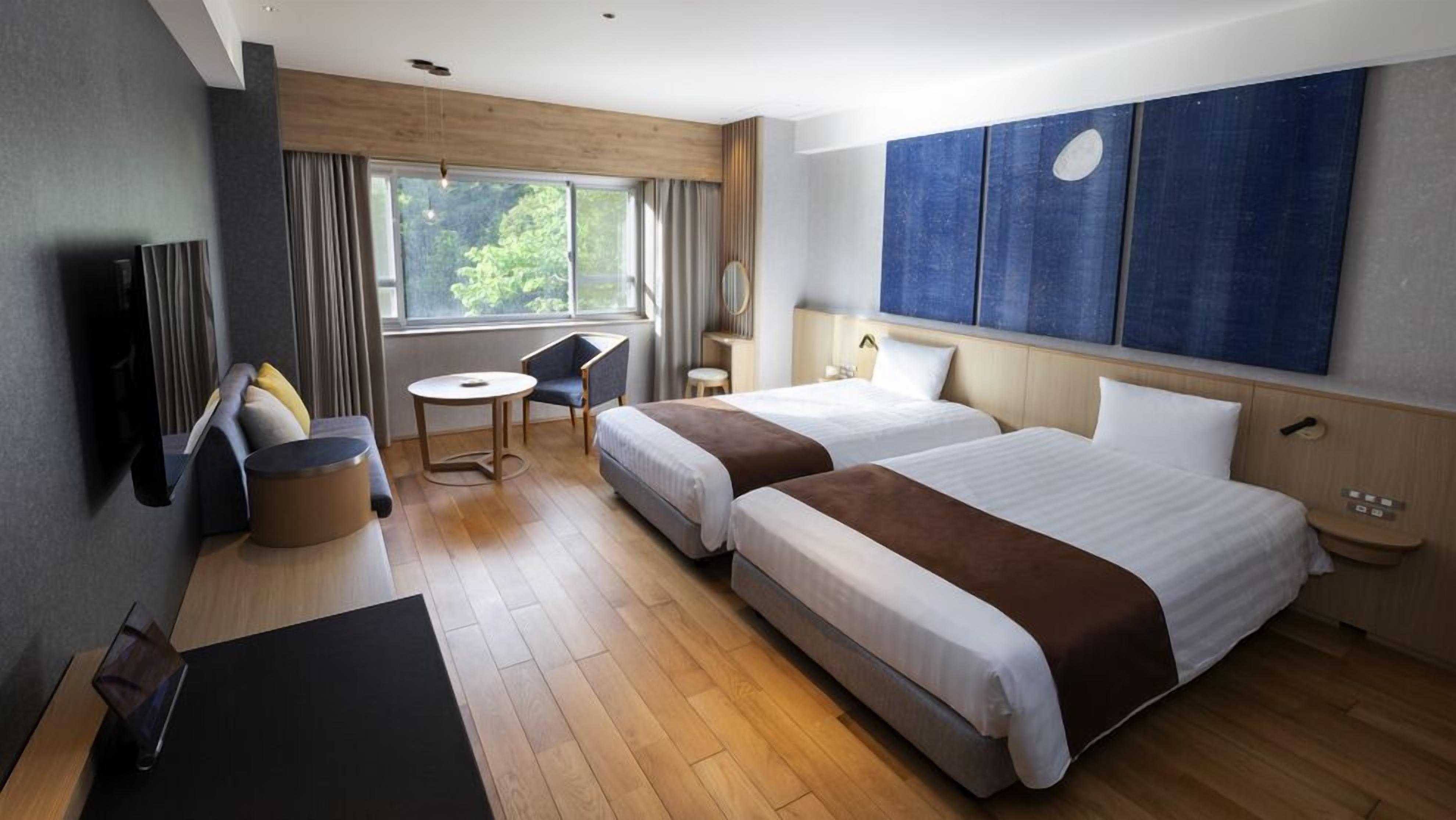 A hotel room with two beds, a desk, a chair, a small table, a TV, and a large window.