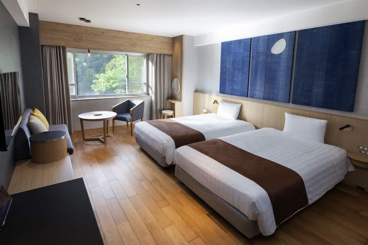 A hotel room with two beds, a desk, a chair, a small table, a TV, and a large window.