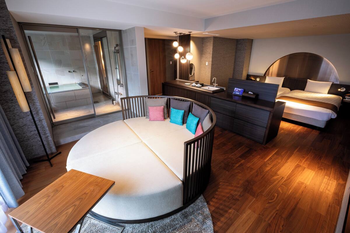 A modern hotel room with a circular bed, a glass shower enclosure, a vanity with a mirror, and a bedside table.