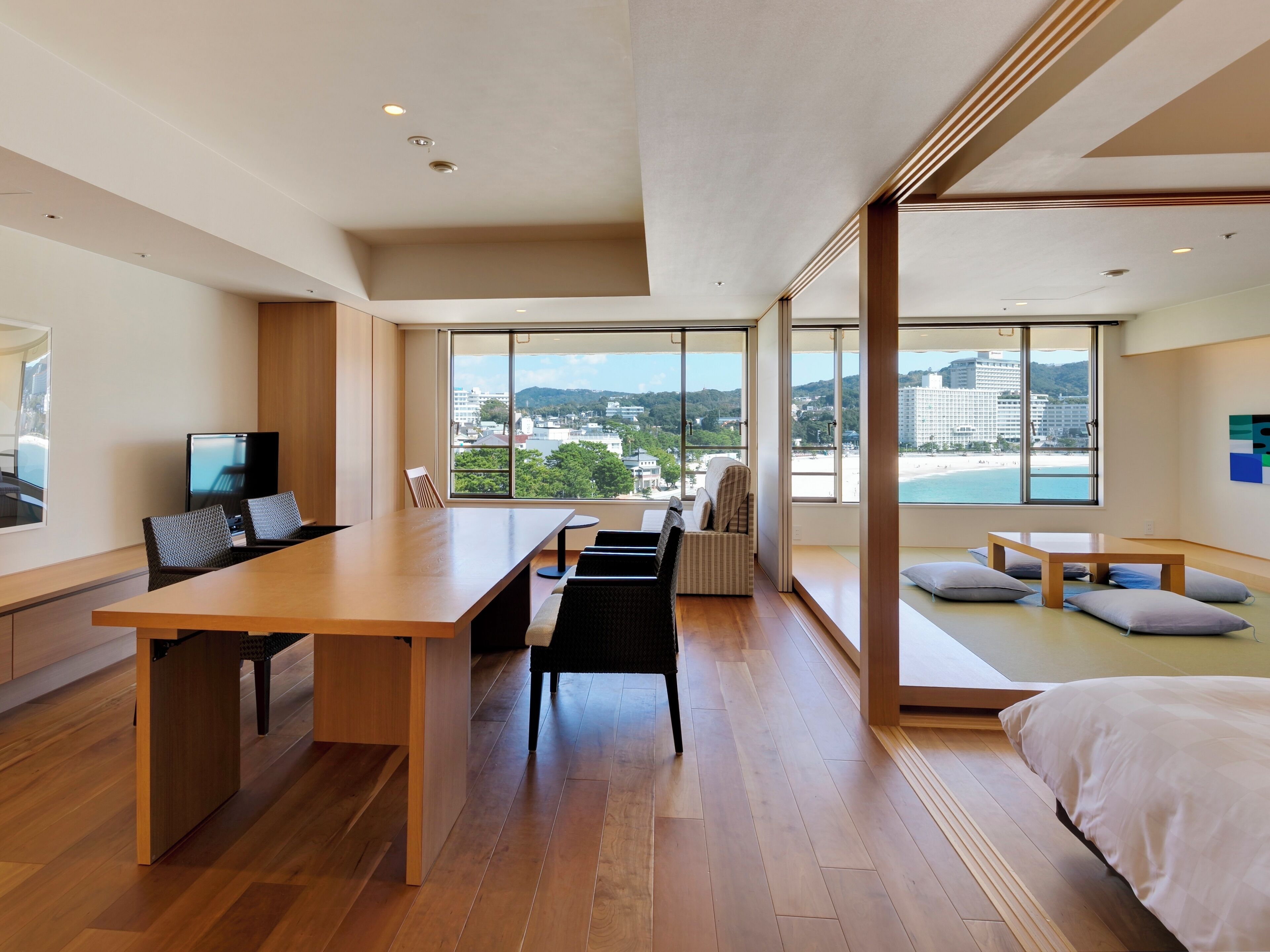A modern hotel room with a large wooden desk, a comfortable seating area with cushions, and large windows offering a view of the city and beach.