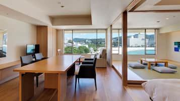 Une chambre d’hôtel moderne dotée d’un grand bureau en bois, d’un coin salon confortable avec des coussins et de grandes fenêtres offrant une vue sur la ville et la plage.