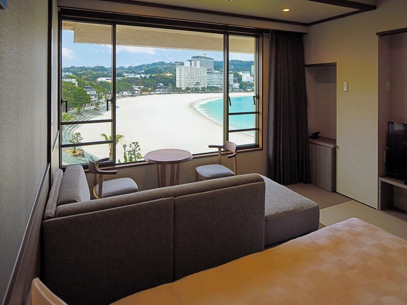 Habitación de hotel con un ventanal amplio que ofrece vistas a una playa y edificios, un sofá, una mesita y un televisor.