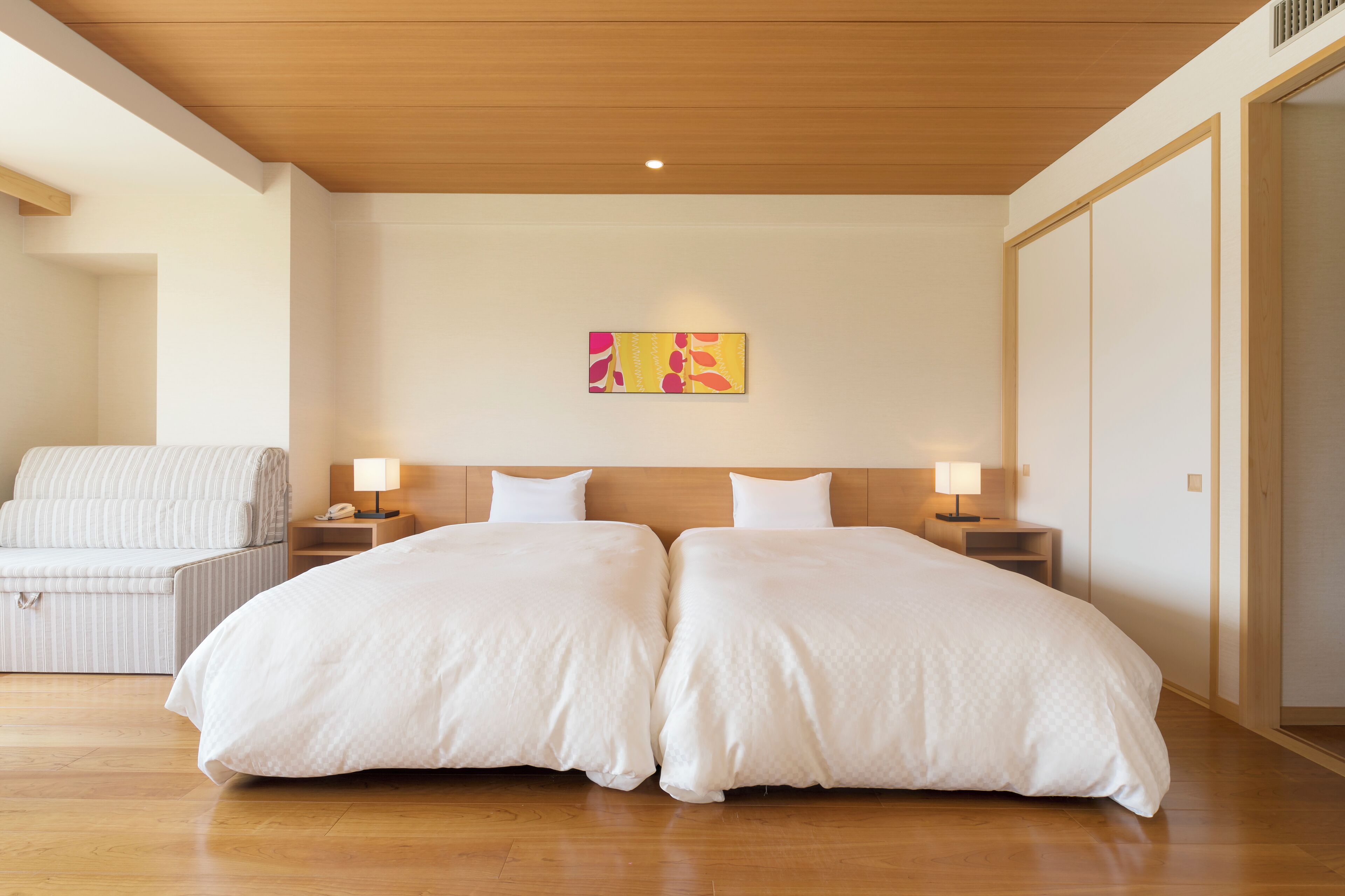 Een hotelkamer met twee bedden, een nachtkastje met lamp, een stoel en een kunstwerk aan de muur.