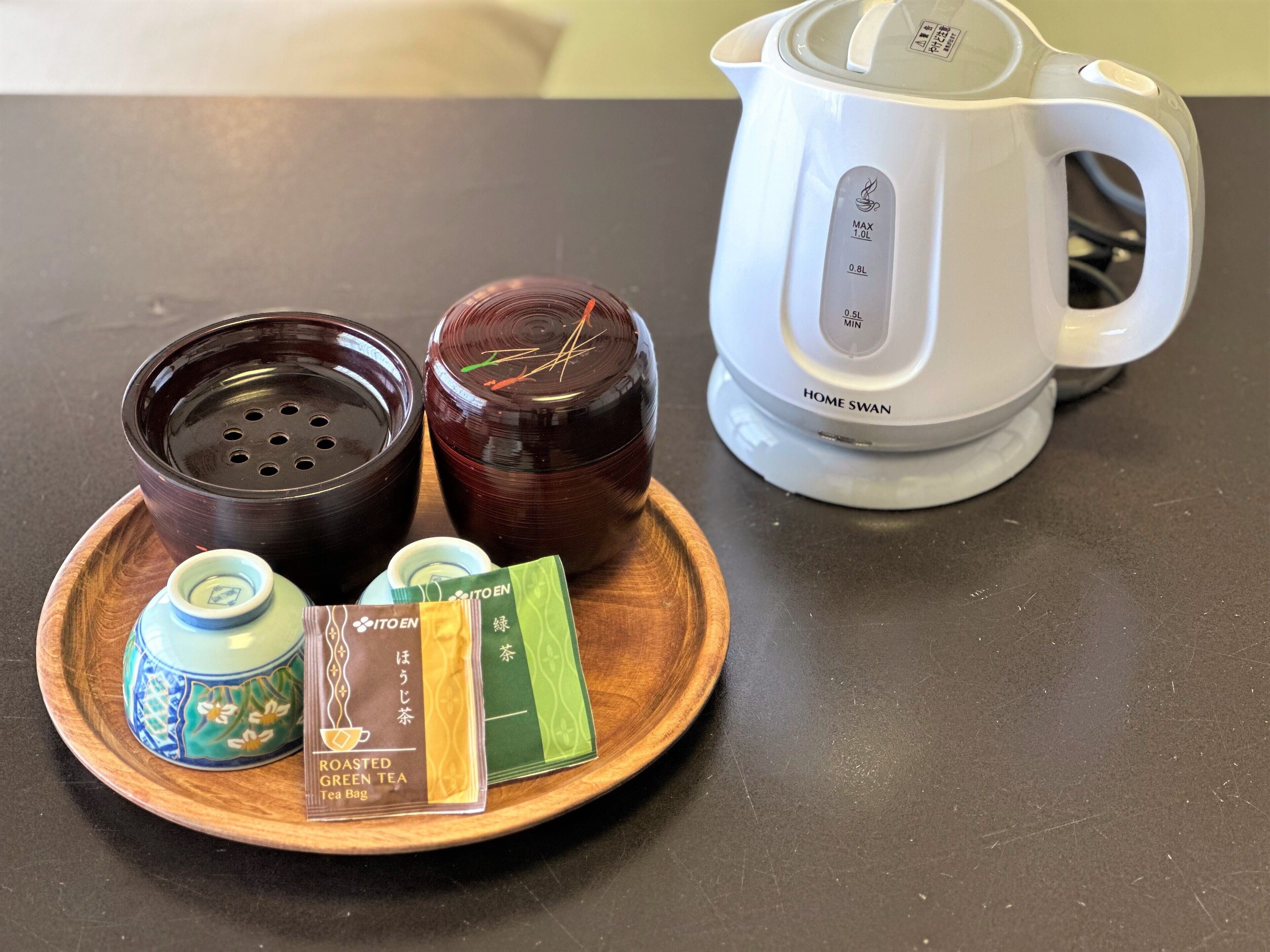 A kitchen counter with a white electric kettle labeled "HOME SWAN," two dark ceramic containers, and a wooden tray holding a tea bag and a tea bag wrapper.