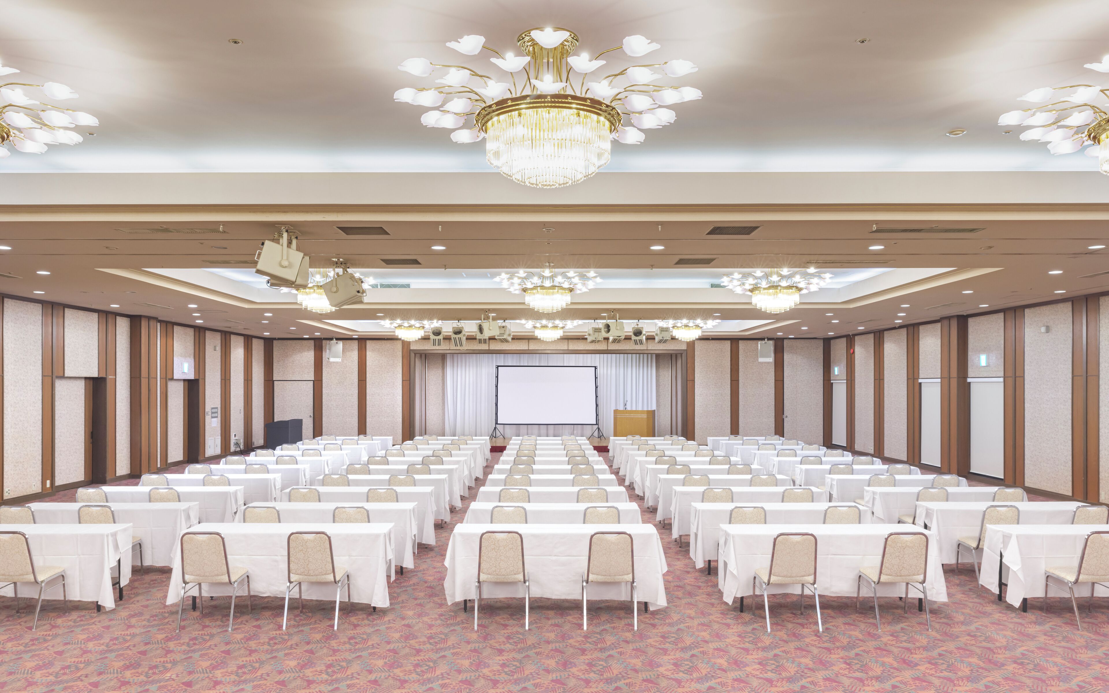 Un amplio salón de conferencias con filas de mesas y sillas, un escenario con proyector y varios candelabros.